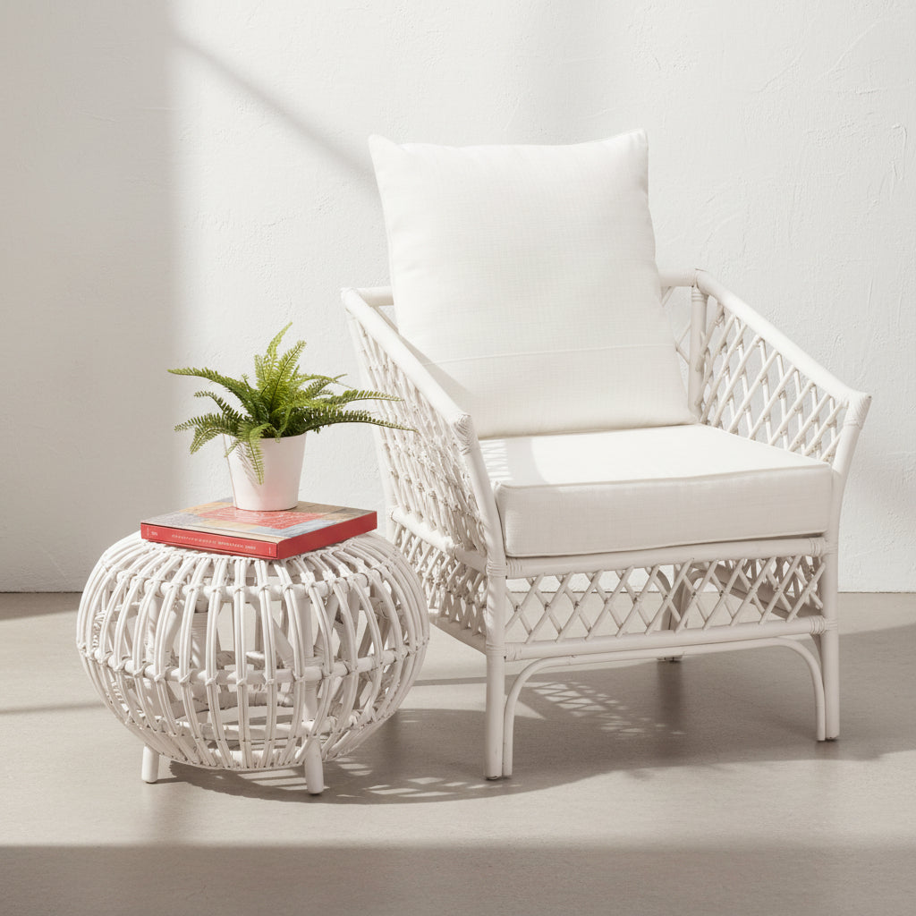 A white rattan armchair with a cushion, placed next to a matching round side table with a plant on top.